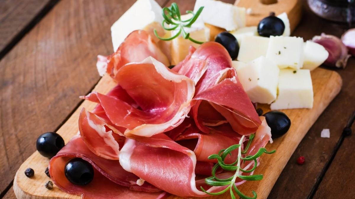    На деревянной доске слайсы хамона, нарезанный кубиком козий сыр, маслины и свежий розмарин:Freepik