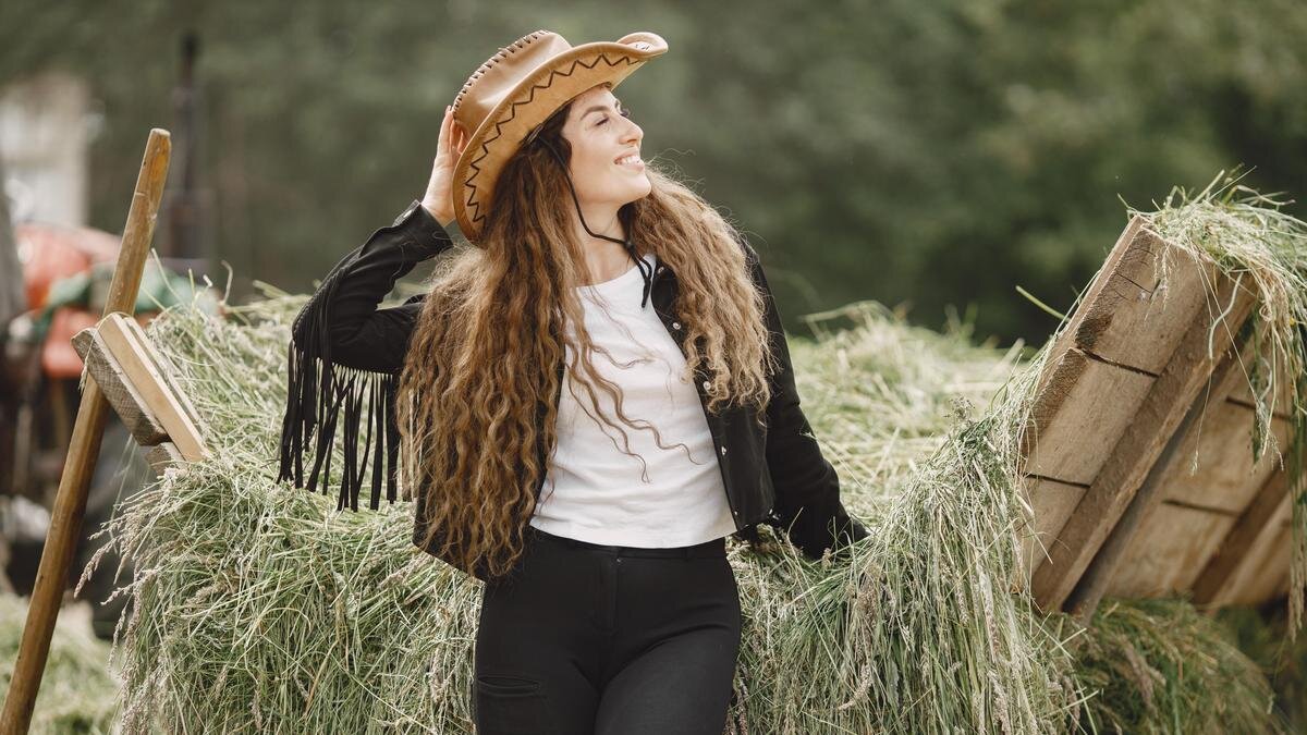    Девушка в брюках, футболке, куртке с бахромой и шляпе стоит около воза с сеном:Freepik