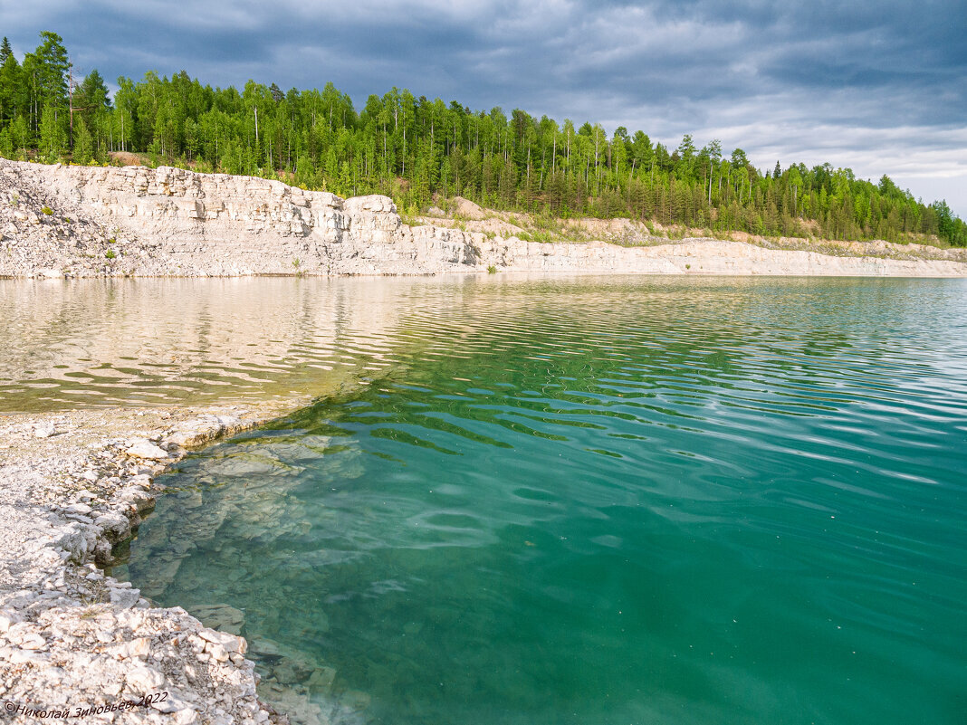 Затопленная часть Бельгопского карьера с водой удивительно красивого морского цвета, как бы дополняет общий антураж с окаменелыми ракушками и кустами девонских кораллов. Обратите внимание на срез гигантских толщей известняка, которые не так глубоко скрывает тайга