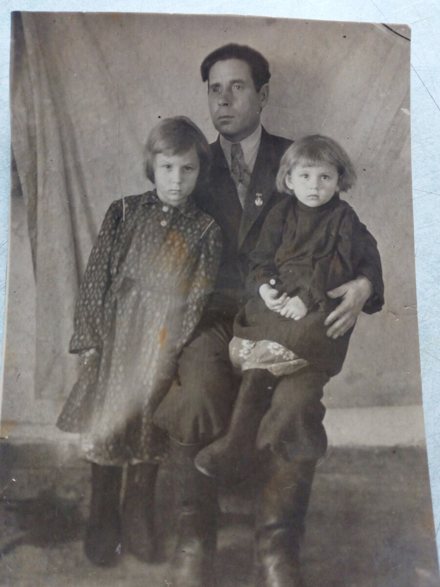 Это я и Аня с отцом. Я у отца на коленях. Не знаю, сколько мне лет. Похоже, что года три. Мы в валенках. значит уже зима. Но помню, что в домике этом тогда почти что гулял ветер. У отца такие большие резиновые сапоги, которые он носил с толстыми носками и портянками, когда ходил по фермам. . 