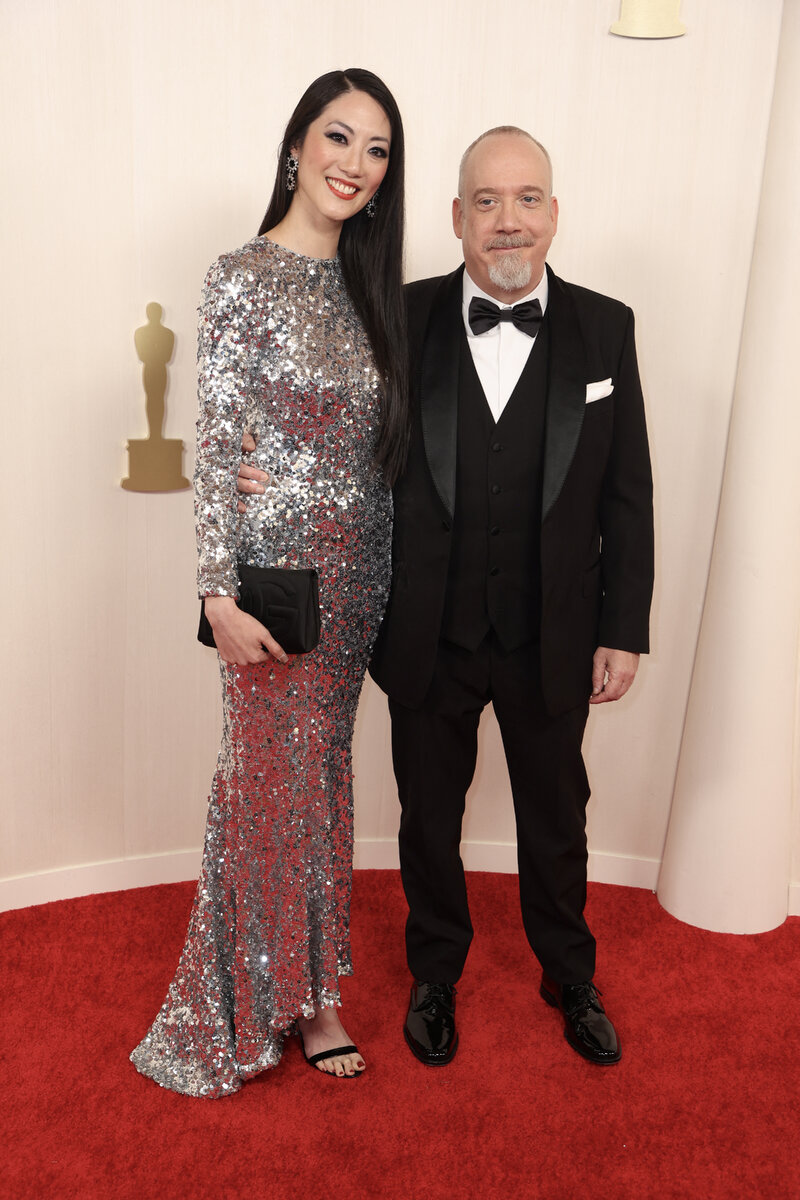 Клара Вонг в Dolce & Gabbana и Пол Джаматти. Лаконичное по фасону платье с длинными рукавами и небольшим эффектом маллет (заднее полотнище длиннее переднего) щедро усыпано мелкими паейтками, которые уравновешены черным сдержанным клатчем и простями черными босоножками.