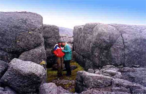 Фаллический камень по форме напоминает знаменитый - Омфал, Пуп Земли, находящийся в Дельфах, известнейшем центре культуры Античного мира. "Греческий" Омфал небольшого (около метра) размера и украшен резным узором по нижней части поверхности. "Кольский Пуп", в отличие от него, крупнее и сильно обветрен. Вместо резьбы, с восточной стороны он имеет вытесанный широкий и плоский "нос" и, образованным вследствие этого "лицом" смотрит на восток, встречая восходящее Солнце. На краю плато перед идолом, расположено подковообразное скопление камней, из которых три больших камня, два по бокам прижатые к кромке обрыва и один посередине отодвинутый вглубь плато образуют отдельную изолированную площадку, идентифицированную нами как жертвенник. Местность вокруг Святилища неординарна, и носит мистический оттенок, подчеркнутый со стороны от юга через запад до севера громадой застилающего горизонт каменного плато, разбитого трещинами безводного плато, похожего на карстовые плато Крыма. С левой стороны края плато его подпирают причудливые каменные изваяния фантастических человекоподобных существ. С правой сплошная пропасть с провалом щели - перевала. Внизу же, за кромкой обрыва, особенно хорошо с отделенного от остального плато жертвенного места, открывается удивительно волнующий вид на широкую каменистую долину Северного цирка Юдычвумчорра, окаймленную, словно чаша отрогами гор Часначорр и Петрелиус. В восточном направлении, внизу, под Петрелиусом, блестит чарующей и завораживающей красотой каскад изумительных бирюзовых озер. Во всем окружающем чувствуется удивительная торжественность и суровая гармония, первозданная красота поднебесного мира, окаймленная клубящимися разводами белых облаков, медленно проплывающих рядом в синеве неба. Я с волнением рассматривал "Кольский Пуп", и из глубин памяти всплывали сведения, прочитанные когда-то о его Древнегреческом собрате - Омфале, о Дельфах, Аполлоне и Гиперборее.