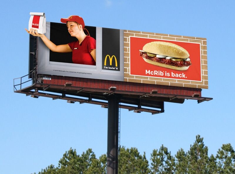 Это рекламный щит McDonald's с увеличенным изображением сотрудника McDonald's, протягивающего зрителям набор McRib. Рядом с этим находится заметное изображение сэндвича McRib с надписью "McRib возвращается" на фирменном красном фоне бренда. Слоган компании "Мне это нравится" также размещен на рекламном щите. 
