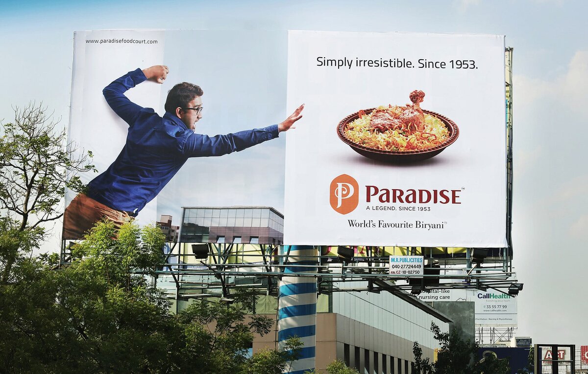 Реклама на рекламном щите подчеркивает давние традиции и популярность ресторана под названием "Paradise", который утверждает, что предлагает "Самое любимое в мире бириани". Привлекательность блюда подчеркивается слоганом "Просто неотразимо. С 1953 года", что говорит о том, что ресторан был любимым кулинарным направлением на протяжении десятилетий. Изображение человека, который, казалось бы, движется к блюду, добавляет динамический элемент, предполагая энтузиазм и желание поесть. 