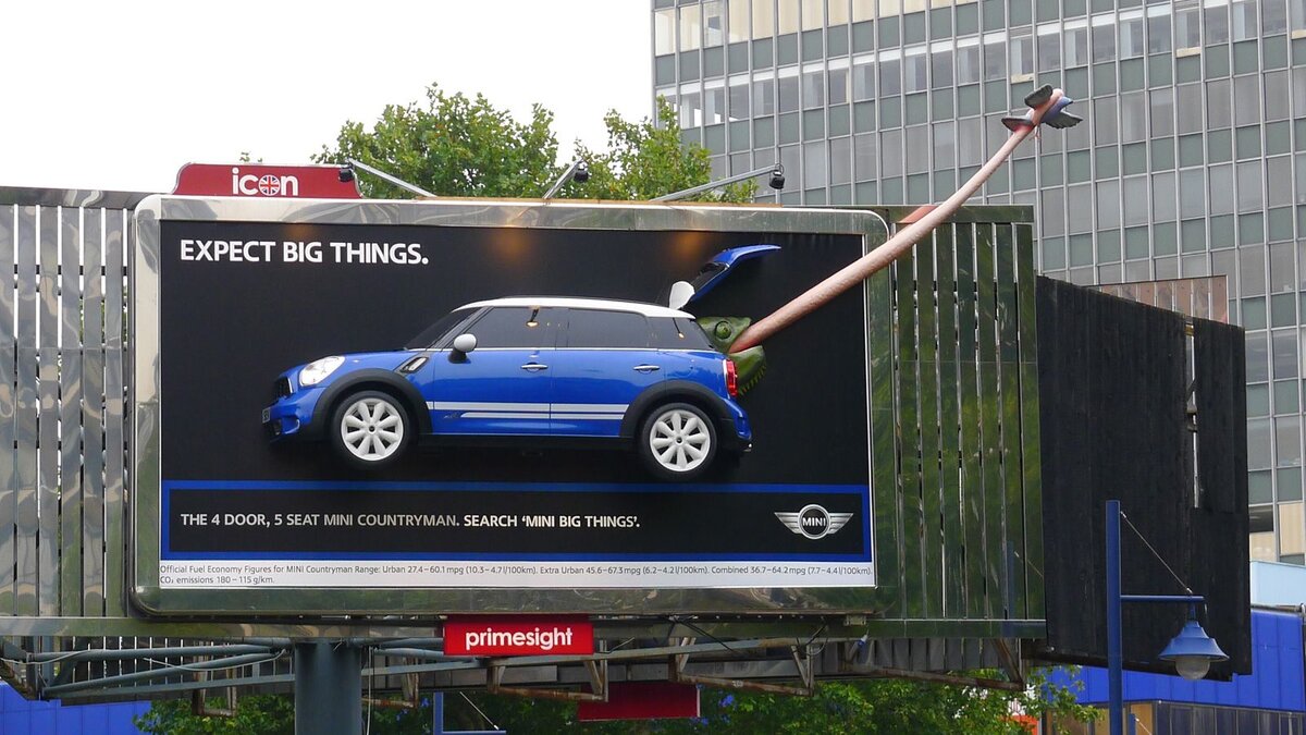 Эта наружная реклама демонстрирует креативный подход, превращая автомобиль MINI Countryman в огромный, неожиданный объект на билборде. Таким образом, она подчеркивает концепцию "Expect Big Things" (Ожидайте большего), буквально показывая маленький автомобиль в увеличенном виде, что играет на контрасте между названием бренда MINI и идеей крупномасштабности. Использование яркого крюка и части реалистичного динозавра, кажется выходящего из багажника, добавляет элемент неожиданности и игривости, привлекая внимание и делая рекламу запоминающейся. 