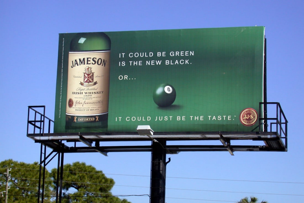 На билборде рекламируется ирландский виски Jameson с лозунгом: "Зелёный — это новый чёрный. Или это просто может быть вкус." Изображение показывает бутылку виски и шар для бильярда под номером 8 на зелёном фоне. 