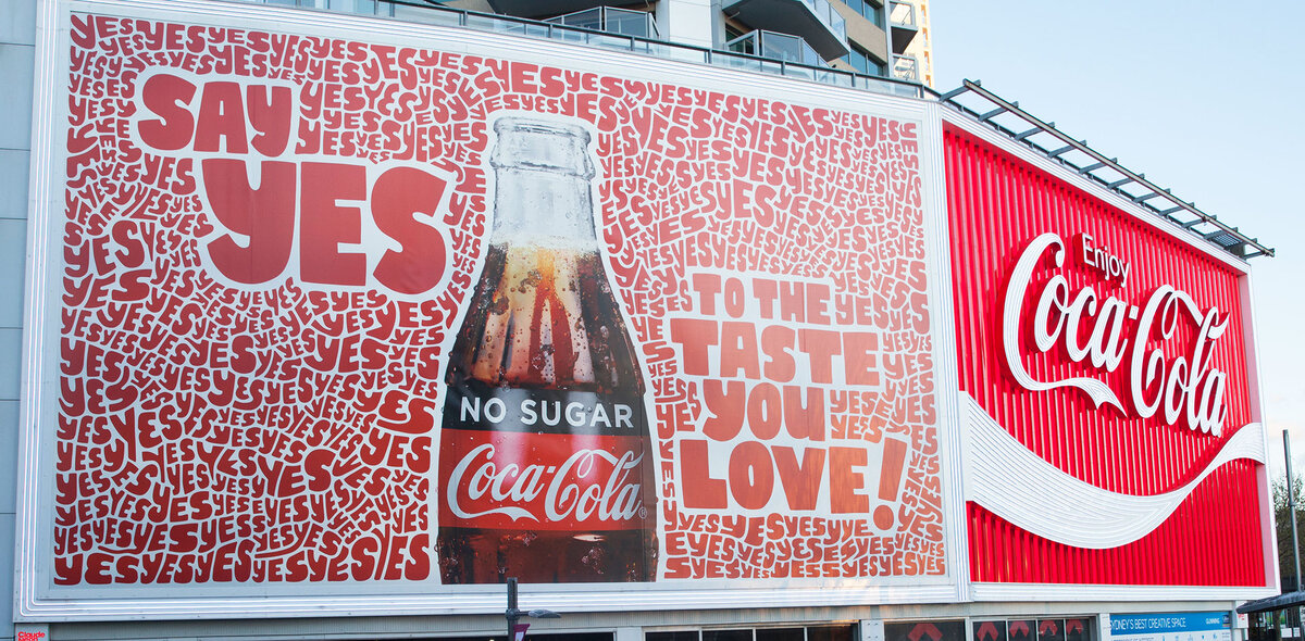 Этот рекламный щит Coca-Cola эффективно привлекает внимание благодаря смелой и увлекательной кампании "Скажи "Да"", использующей повторяющиеся позитивные сообщения и культовые образы бренда. Ярко-красная цветовая гамма и узнаваемый логотип Coca-Cola еще больше повышают узнаваемость бренда и привлекают взгляды зрителей. 