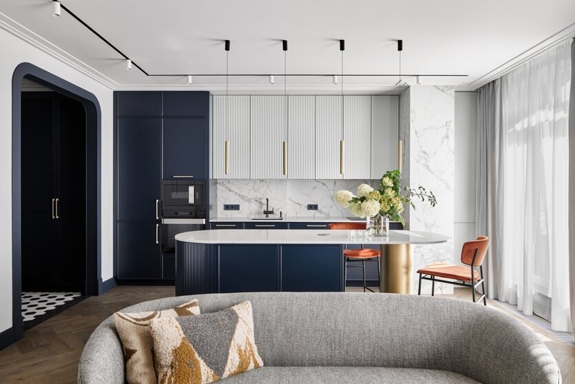 Вид из гостиной на кухню-столовую. Кухня оснащена системами хранения и радиусным островом с обеденной зоной и секциями для хранения. Светильники, «Центрсвет».