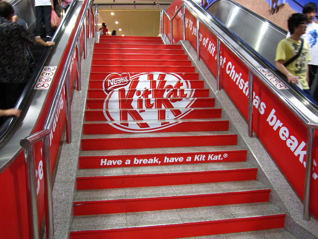 Эта реклама KitKat эффективно привлекает внимание потребителей, используя ступени эскалатора как креативный холст для слогана "Have a break, have a Kit Kat," что напоминает о возможности отдыха с шоколадкой во время повседневной суеты. 
