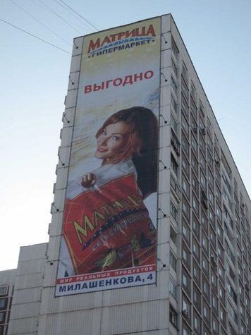 На здании размещен большой рекламный баннер гипермаркета "Матрица" с изображением улыбающейся женщины и надписью "Выгодно" на фоне голубого неба. 
