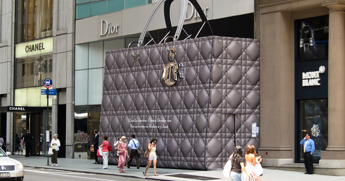 На улице размещен гигантский рекламный щит в виде сумки Dior, привлекающий внимание прохожих и анонсирующий открытие нового бутика. 