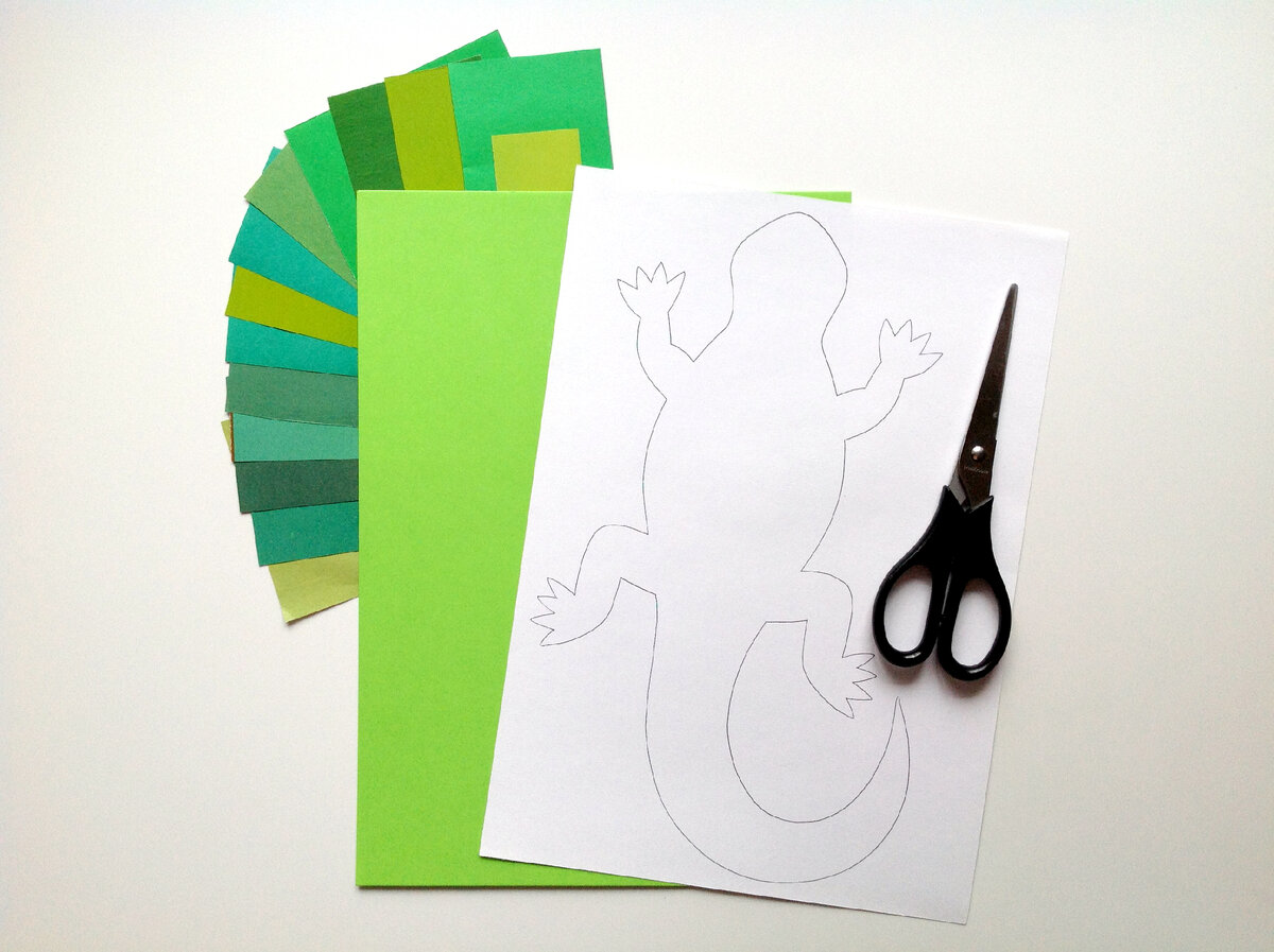 Выбираем цветной картон для фона. Можно представить, что ящерица находится в траве, на  песке или камнях, в зависимости от придуманного пространства подберите картон.  Выберите картон контрастного цвета по отношению к цвету ящерицы, чтобы она лучше читалась на фоне. Я выбрала зеленый цвет картона для своей аппликации, чтобы создать вокруг ящерицы лужайку с травой.  На белом листе бумаги простым карандашом рисуем силуэт ящерицы.  Так как моя ящерица будет в траве, для листиков и травинок я подобрала кусочки бумаги  разных оттенков зеленого. 
