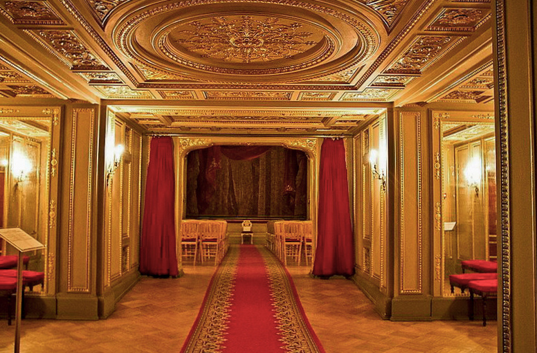 Их приветствует уютный аванзал с роскошным лепным потолком, идеальными зеркалами и изысканными канделябрами, мягко мерцающими тёплым светом. Театралы проходят через золочёный портал и попадают в зрительный зал с партером и двумя ярусами. Фотография с просторов интернета, цветокоррекция автора статьи. 