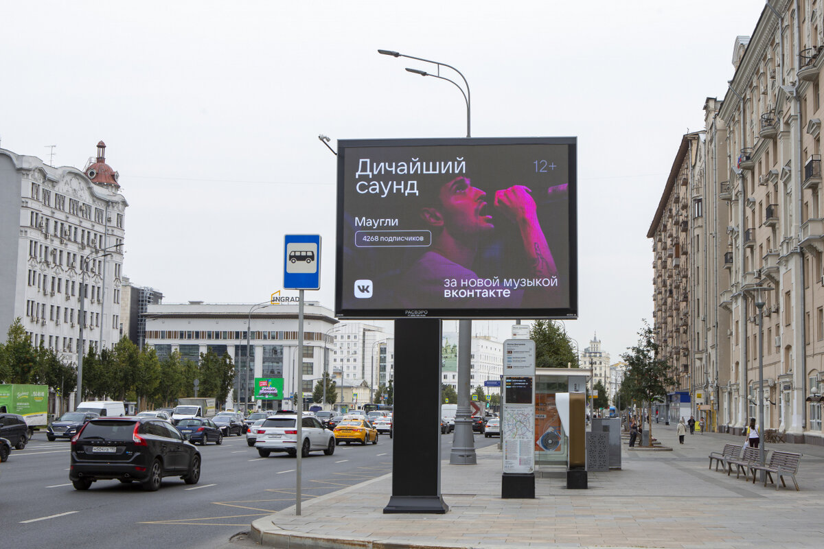 Эффективная наружная реклама музыкального концерта, отображенная на цифровом билборде в оживленном районе города, привлекает внимание ярким изображением исполнителя и информацией о мероприятии, включая количество подписчиков и платформу для прослушивания. 