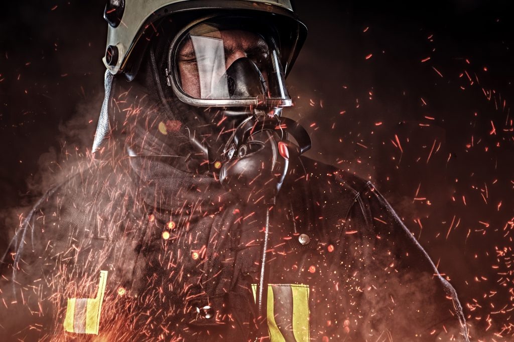    A professional firefighter dressed in uniform and an oxygen mask standing in fire sparks and smoke over a dark background. Марк Волков