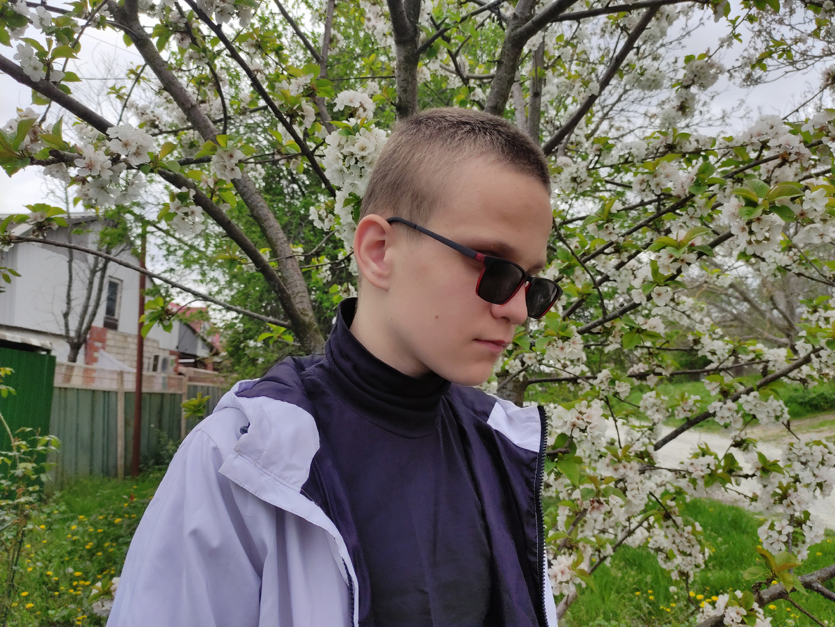 Тифлокомментарий. На фото Гошка. Высокий, коротко стриженный подросток в чёрных очках. Фото сделано весной, на Гоше белая куртка и чёрная водолазка. На заднем фоне цветёт вишня