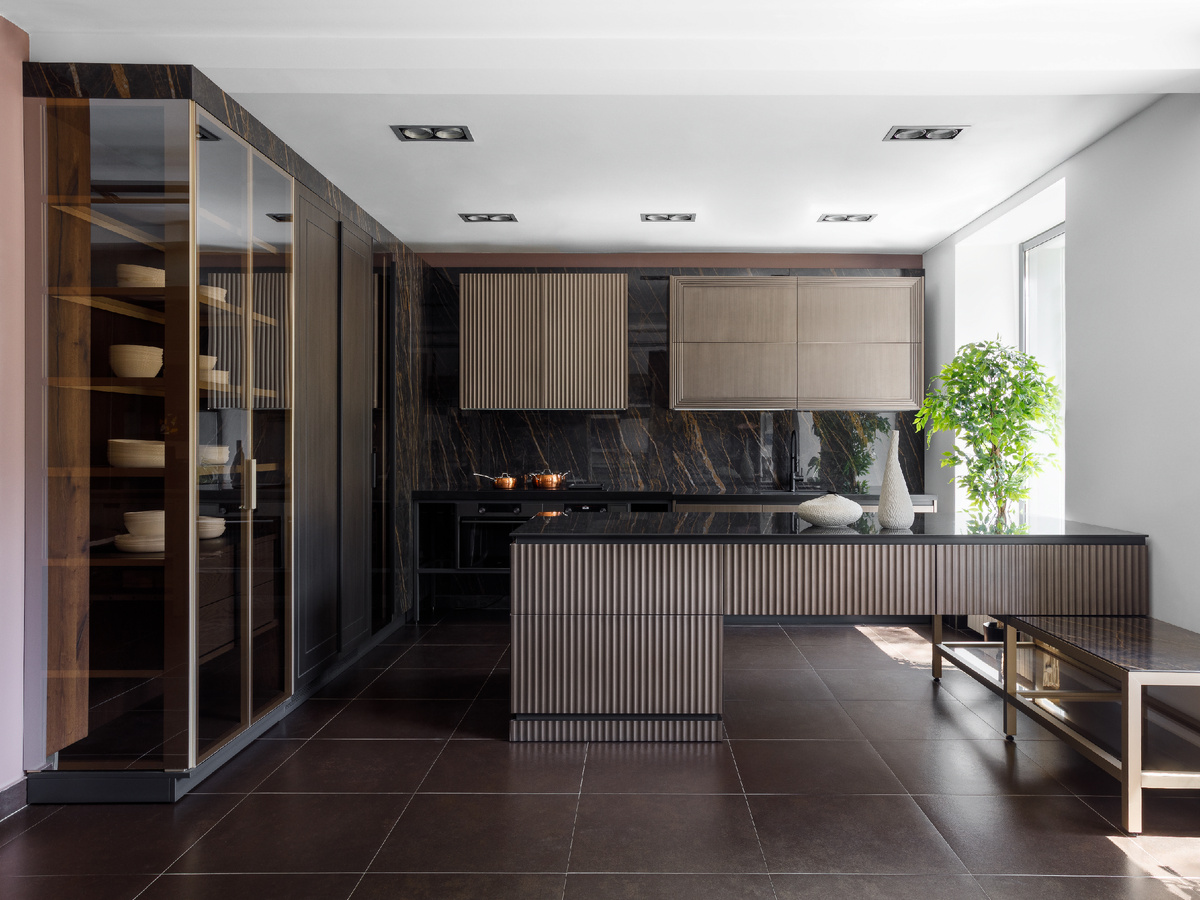 Притягивают внимание и задают характер темные оттенки эмали под бронзу, керамики с рисунком под мрамор, шпона Дуба в винтажной отделке в сочетании со стеклом и алюминиевыми конструкциями. Декоративная подсветка подчеркивает игру объемов, а также внутреннее наполнение витрин, тем самым дополняя разноуровневый сценарий освещения помещения в целом.