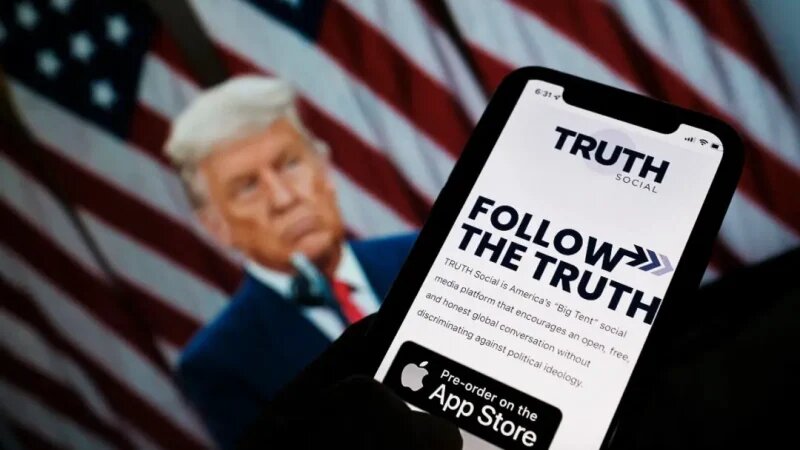 Человек проверяет наличие Truth Social в App Store на смартфоне, на фоне фотографии Дональда Трампа на экране компьютера в Лос-Анджелесе, 20 октября 2021 года. (Chris Delmas/AFP via Getty Images)