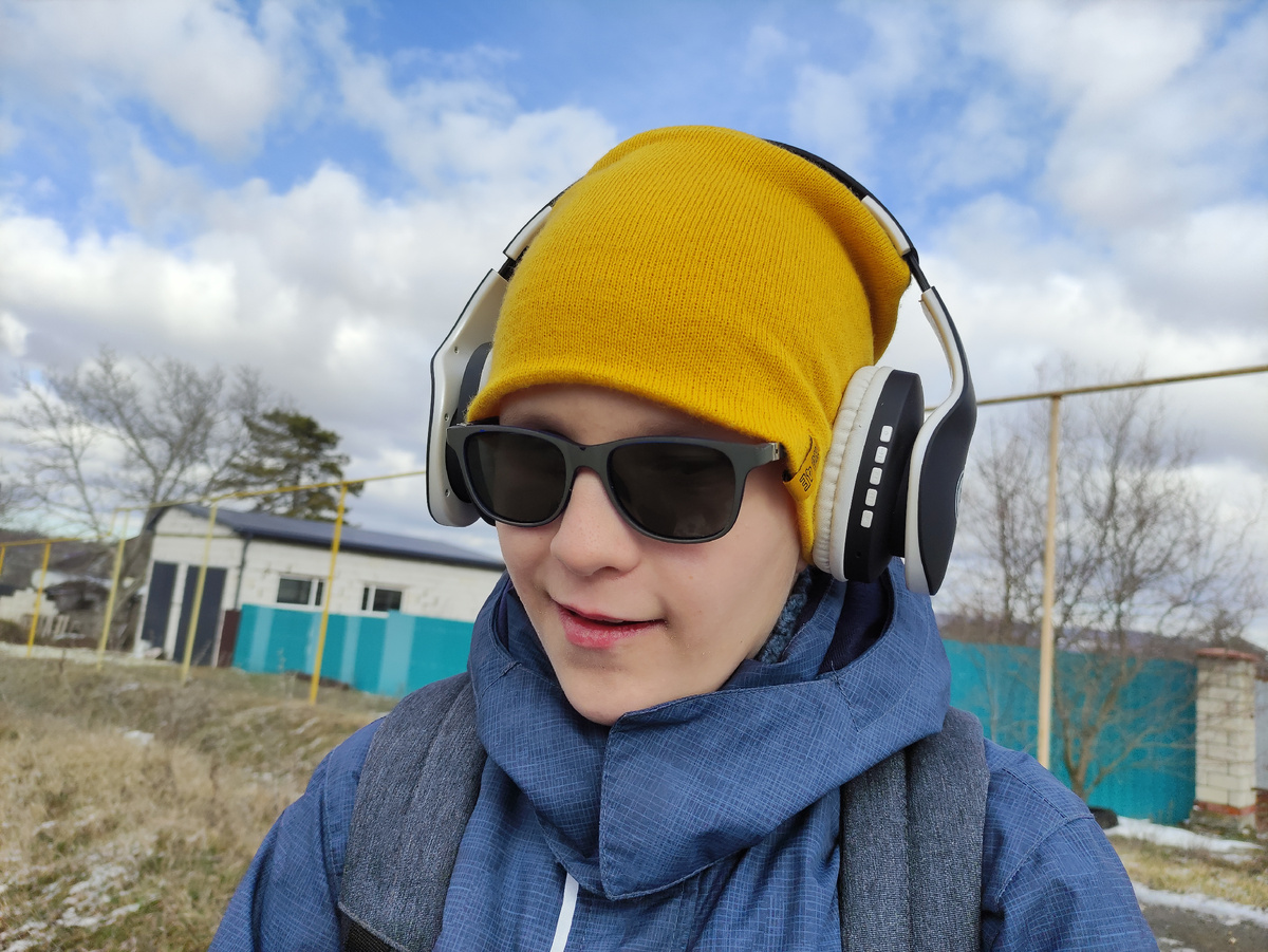 Тифлокомментарий. Подросток в синей куртке и жёлтой шапке на улице. На ушах наушники. Южная зима без снега