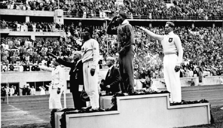   Оуэнс отдает честь американскому флагу после победы в прыжках в длину на летних Олимпийских играх 1936 года. (Слева направо) Наото Тадзима, Оуэнс, Луз Лонг Фото: Bundesarchiv, Bild 183-R96374. Собственная работа, по лицензии CC BY-SA 3.0