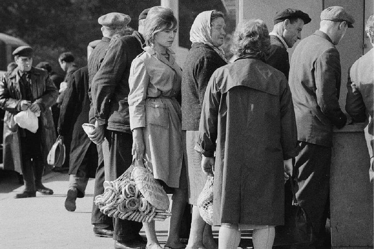 Девушка, стоя в очереди, о чем-то задумалась. В правой руке она уверенно держит типичную авоську, набитую под завязку добычей (хлебо-булочными изделиями) (Фотография времен СССР)