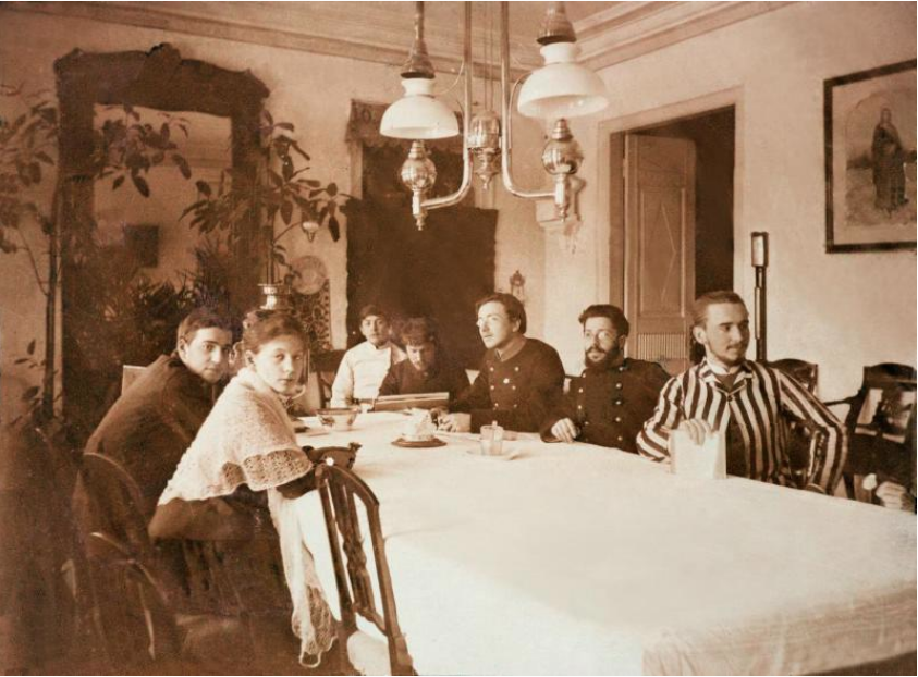 Фотография любительская. В столовой Мамонтовых в Абрамцеве. За столом сидят слева направо: В.С. Мамонтов, Нат. Анат. Мамонтова, неуст. молодой человек, В.А. Серов, неуст. молодой человек в форменном мундире, неуст. лицо и С.Ф. Чиж (молодой мужчина в полосатой куртке). Из собрания Музея-заповедника "Абрамцево"