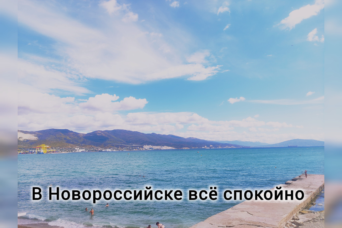 Тифлокомментарий. Снимок Цемесской бухты с набережной Новороссийска. За бухтой Маркхотский хребет. Сверху голубое небо с облаками. Текст: в Новороссийске всё спокойно 