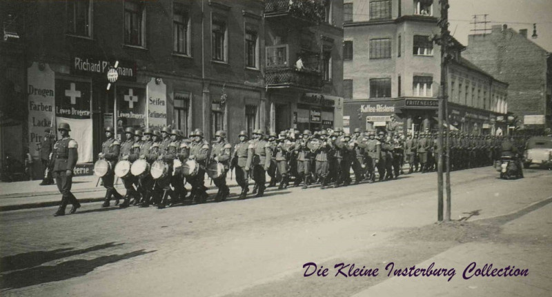 Оркестр 43-го пехотного полка следует по южной части Гинденбургштрассе (бывшей Банхофштрассе) в сторону железнодорожного вокзала, через перекрёсток с Луизенштрассе. Конец 30-х/начало 40-х годов XX века. На фотографии можно видеть парфюмерно-галантерейный магазин Рихарда Плауманна, а также магазин колониальных и хозяйственных товаров Фрица Нельсона. Фотограф стоит спиной к дому автора данной статьи, Вильгельма Замейтата. Фото из собрания фонда "Die Kleine Insterburg" Евгения А. Стюарта.
