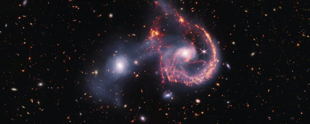    This composite image of Arp 107, created with data from the James Webb Space Telescope’s NIRCam (Near-InfraRed Camera) and MIRI (Mid-InfraRed Instrument), reveals a wealth of information about the star formation taking place in these two galaxies and how they collided hundreds of million years ago. The near-infrared data, shown in white, show older stars, which shine brightly in both galaxies, as well as the tenuous gas bridge that runs between them. The vibrant background galaxies are also brightly illuminated at these wavelengths. On the other hand, MIRI data show the young stars and star-forming regions in vibrant orange and red. Our view in the mid-infrared provides the best view of the collision point, given the noticeable gap at the top of the spiral galaxy. This collision not only began a new bout of star formation in the region, but also produced an endearing smile. [Image description: A pair of interacting galaxies. The larger of the two galaxies is slightly right of centre, and is composed of a hazy, bright, white centre and a ring of gaseous filaments, which are different shades of red and orange. Toward the bottom left and bottom right of the ring are filaments of gas spiralling inward toward the core. At the top left of the ring is a noticeable gap, bordered by two large, orange pockets of dust and gas. The smaller galaxy is made of hazy and white gas and dust, which become more diffuse further away from its centre. To this galaxy’s bottom left, there is a smaller, more diffuse gas cloud that wafts outward toward the edges of the image. Many red, orange, and white galaxies are spread throughout, with some being hazier in appearance and others having more defined spiral patterns.] Александр Шереметьев