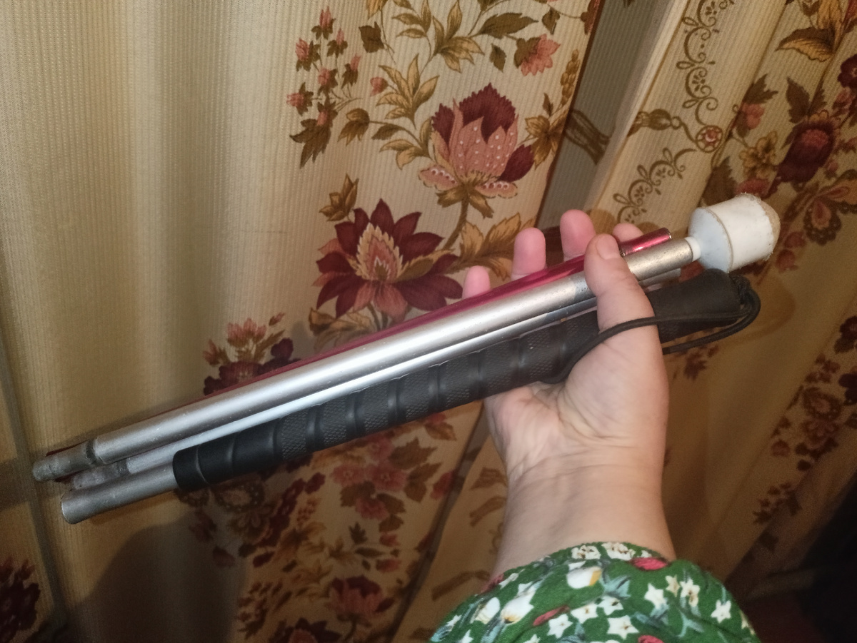 Моя белая трость из четырёх секций, собранная, в правой руке, на фоне светлых штор с цветами. 