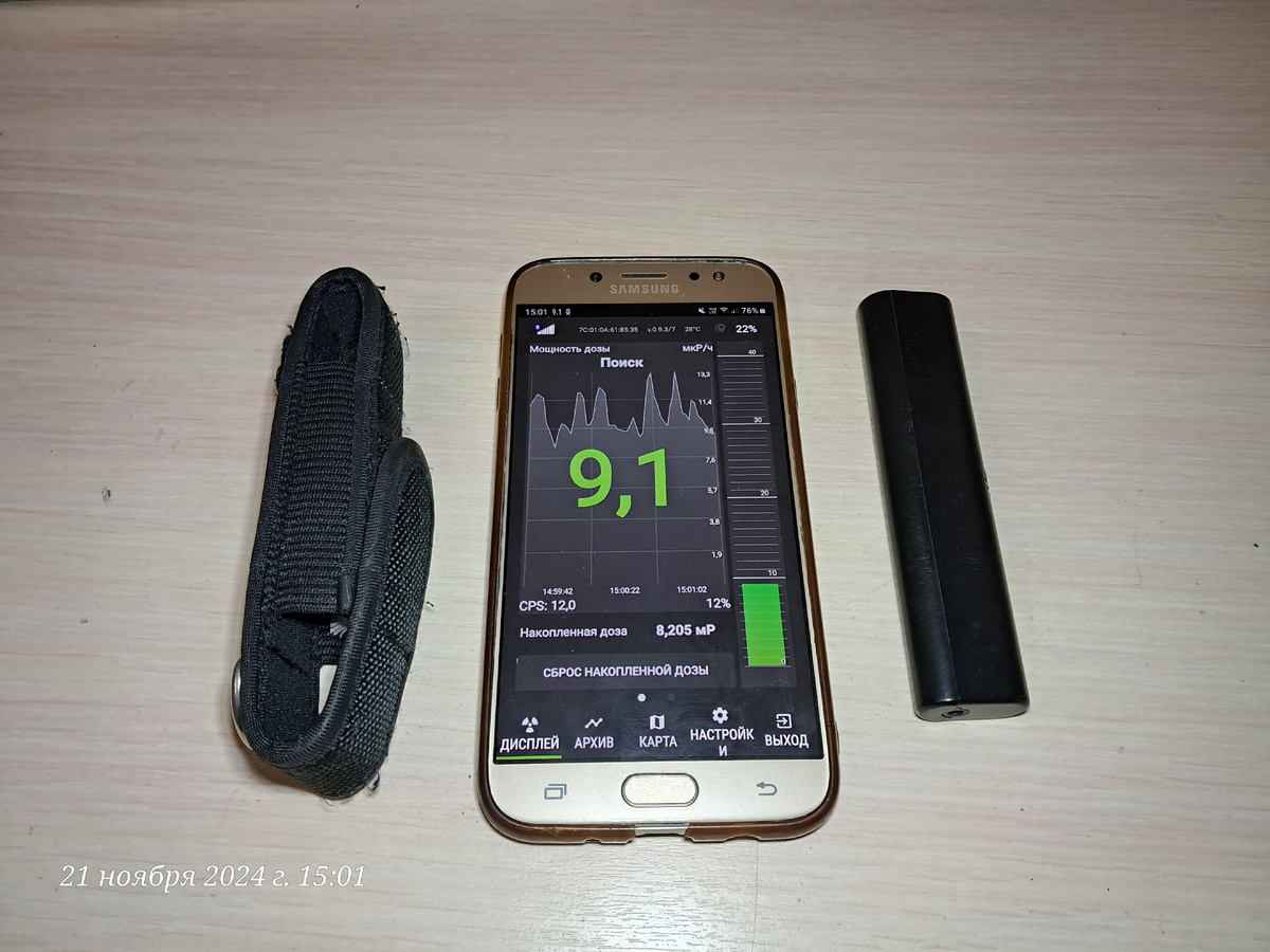 Справа от смартфона дозиметр-радиометр Атом Фаст 8850, слева от смартфона его потрёпанный чехол. На самом смартфоне работает приложение Atom Swift на котором каждые 2 секунды отображаются статистические данные окружающего радиационного фона.