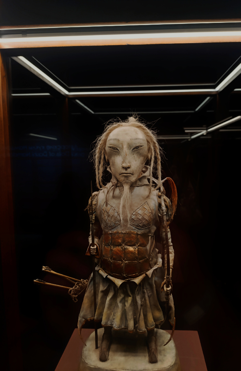 Кукла "Воин" в древних доспехах, с луком и копьём в руках. Лучник спокоен, но готов к атаке. Его причёска соответствует тогдашней моде: бритая макушка и длинные косички, всё это символ непобедимости. Подошва с загнутыми к верху носиками, по обычаям, не способна потревожить землю-кормилицу.