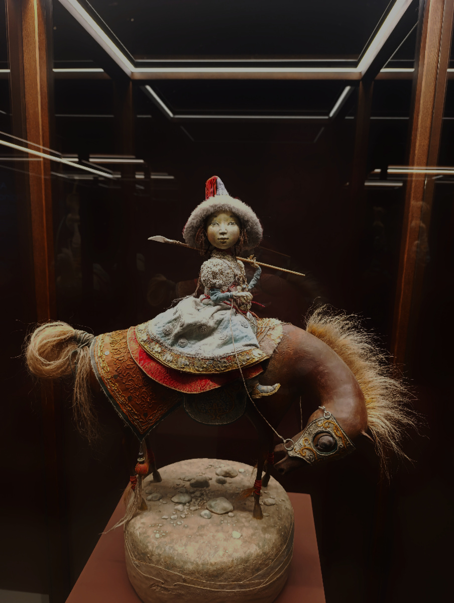 Кукла "Всадница". Героиня древних бурятских сказаниях о девушках-воительницах, совершавших подвиги ради спасения своих братьев. Несмотря на копье в руках, всадница кажется милой и хрупкой. Здесь очень много деталей одежды: национальное платье, сапожки, ожерелье, шапка с меховым оборотом. А лотос на седле имеет особое значение - как символ юности, невинности, чистоты помыслов. 