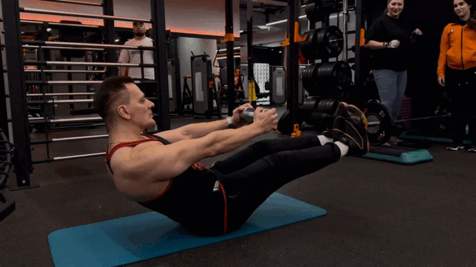 Сядьте на пол, руки с весом вытяните вперёд, а затем поднимите ноги над землей. Ноги должны быть прямые в коленях или немного согнуты