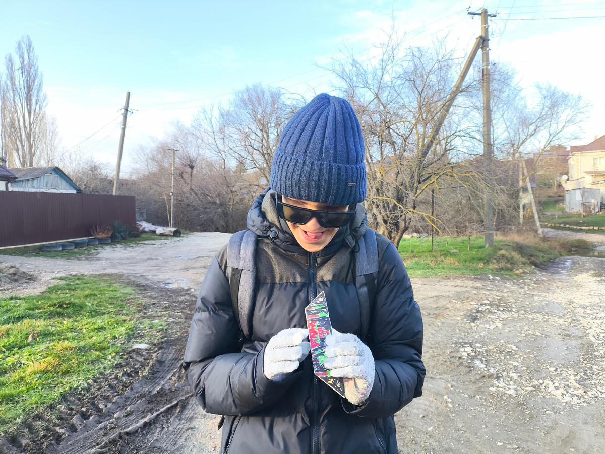 Тифлокомментарий. Гоша, подросток в синей куртке и синей шапке держит в руках бумажный кораблик. Гоша улыбается. 