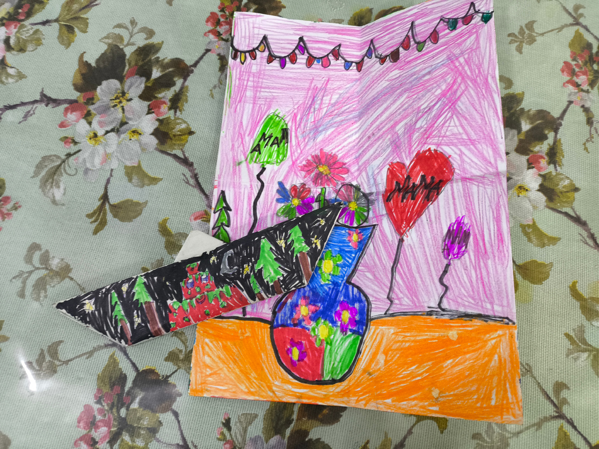 Тифлокомментарий. Открытка, нарисованная фломастерами. Ваза, букет, вокруг шарики, на которых наивно написано "Мама". 