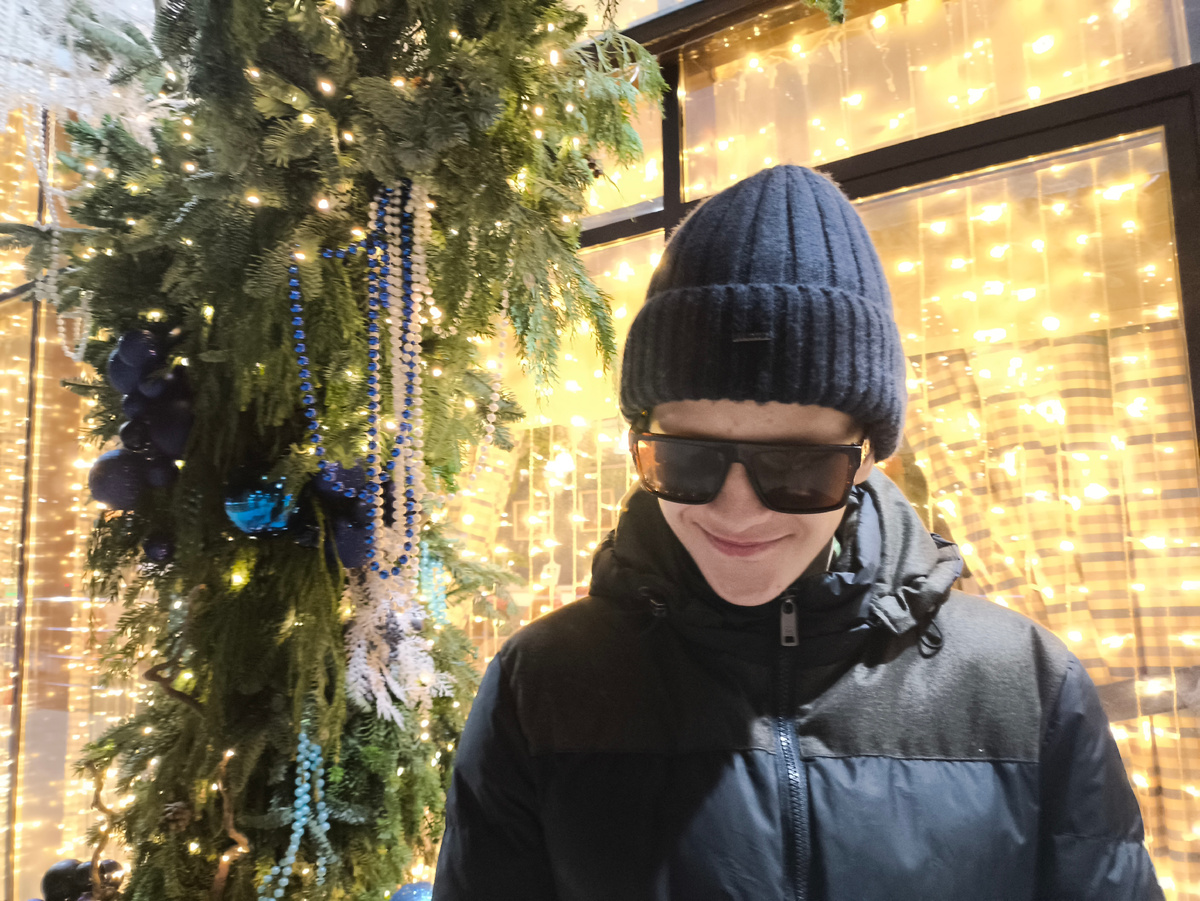 Тифлокомментарий. Подросток в чёрных очках, чёрных шапке и куртке стоит на фоне окна, увешанного золотыми гирляндами. Позади гирлянда из деловых веток