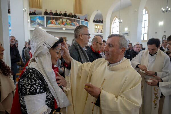 Отец Фран Колай крестит женщину в традиционном албанском наряде в Ллапушнике.Кредит...Арменд Нимани для The New York TimesИзображение