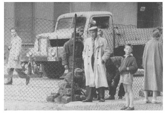 Воскресенье, 13 августа 1961 года. Агитатор из Восточного Берлина в белом пальто и кепке газетчика рычит, пока Ханс-Йоахим Хельвиг-Вильсон фотографирует его у бара Heidelberger Krug. Недавно возведённый проволочный забор был предшественником бетонной стены 