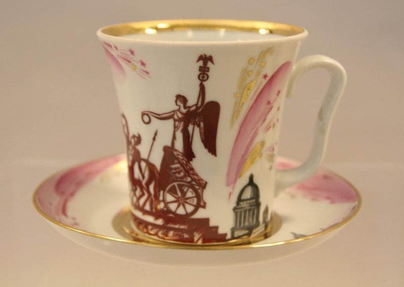 Бокал с блюдцем "Колесница Победы". 1986. Автор формы Криммер Э.М., автор росписи Соколов С.Н. Здесь и далее художник сосредоточился на изображении архитектурных красот города на Неве.  Ручная роспись салюта, исполненная в розовом колере и в золоте, освежают несколько суровый монохромный ряд.Федеральное государственное бюджетное учреждение культуры "Государственный Эрмитаж".