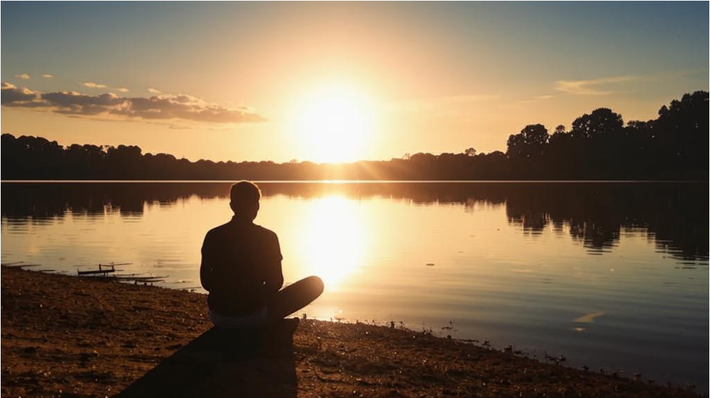 Человек сидит на берегу озера, глядя на отражение солнца в воде, символизируя момент "здесь и сейчас".