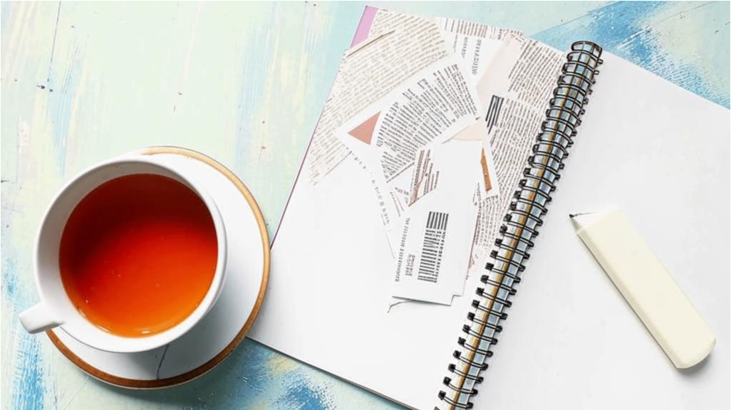 Чашка чая рядом с раскрытой книгой и записной книжкой, в которую кто-то пишет утренние мысли.
