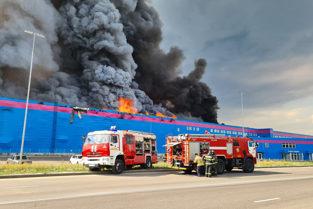 Пожар на складе Ozon в Подмосковье (2022): Огромный черный дым поднимается в небо, языки пламени вырываются из здания. Пожарные машины и спасатели на месте происшествия пытаются справиться с огнем. Пожарные машины и спасатели на месте происшествия пытаются справиться с огнем.