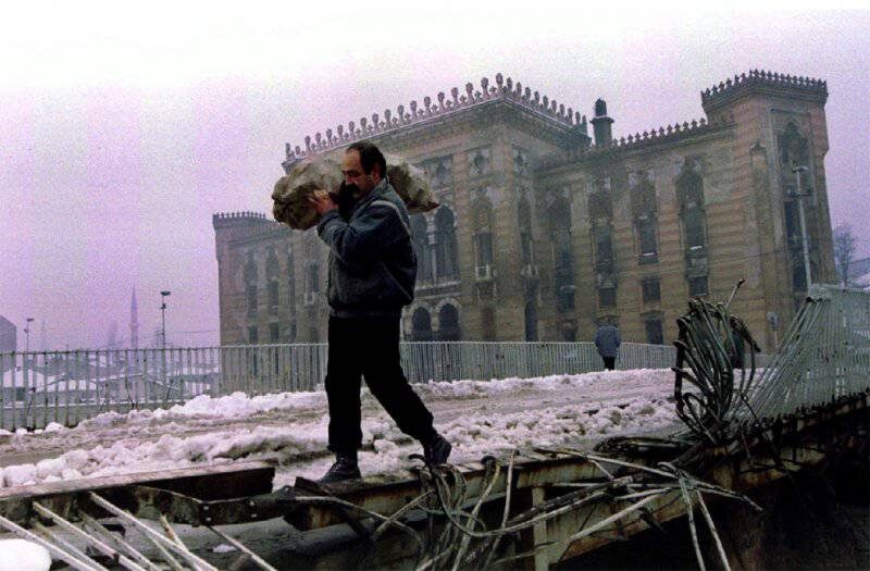 Мужчина несет мешок с дровами по поврежденному мосту. На заднем плане национальная библиотека. Фото из открытых источников.