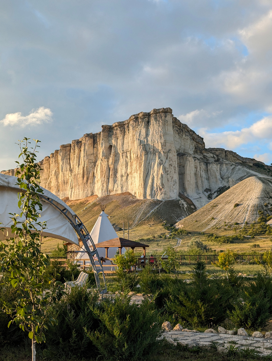 Белая скала Ак-Кая: Крымский "айсберг" посреди степиБелая скала, или Ак-Кая (крымскотат. Aq Qaya, Акъ Къая), – это впечатляющая скалистая гора в Крыму, расположенная возле села Белая Скала Белогорского района. Ак-Кая переводится с крымскотатарского как "белая скала"25. Эта достопримечательность выделяется на фоне крымских пейзажей и привлекает туристов своей необычной красотой156.ОписаниеБелая скала возвышается над долиной реки Биюк-Карасу. Высота скалы составляет 325 метров над уровнем моря, при этом перепад высот между долиной и вершиной достигает 100 метров. Вертикальная стена белого цвета создана из меловых и палеогеновых известняков и песчаников под воздействием эрозии и выветривания. Благодаря этим процессам на вершине образовались столбы, гроты и овальные ниши2.ИсторияАк-Кая – это не только природный памятник, но и место с богатой историей.Древние времена: Здесь обнаружены следы стоянок первобытных людей мустьерской эпохи, а также останки неандертальца и его ребенка346. Предполагается, что здесь жили сарматы и скифы126.Средние века: Белая скала использовалась как место казни.Крымское ханство: Здесь находился главный центр рода Ширин, и на скале избирали старшего рода.В составе России: В 1777 году на Белой скале располагался штаб А. В. Суворова124. В 1783 году князь Потемкин принимал здесь присягу на верность России от крымской знати123.Интересные фактыБелая скала служила местом проведения торжественных сборищ, на которых избирали новых предводителей.В районе Ак-Кая было найдено около 20 стоянок первобытных людей, каменные ножи и орудия труда, а также кости мамонта, пещерного медведя и других животных236.У подножия скалы растет 800-летний дуб, который называют Суворовским.Белая скала – это уникальное место в Крыму, которое сочетает в себе красоту природы и богатую историю. Это место, где можно прикоснуться к прошлому и насладиться величием природы.