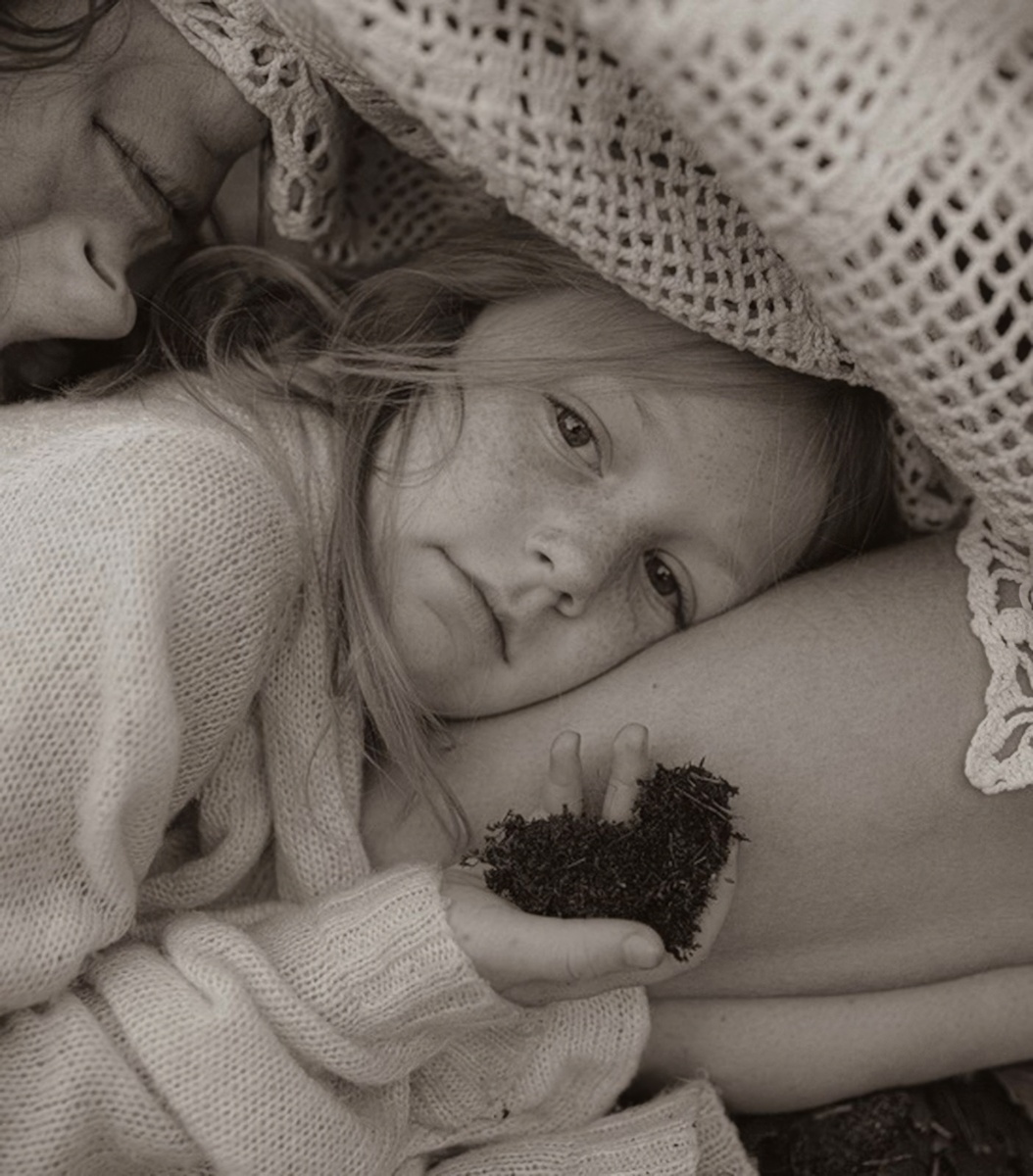 (На фотографии: девочка в свитере прижимается к матери, укрывшись вязаным покрывалом. Ее взгляд пронизывает — одновременно задумчивый и отчужденный. В руке у нее немного земли.)