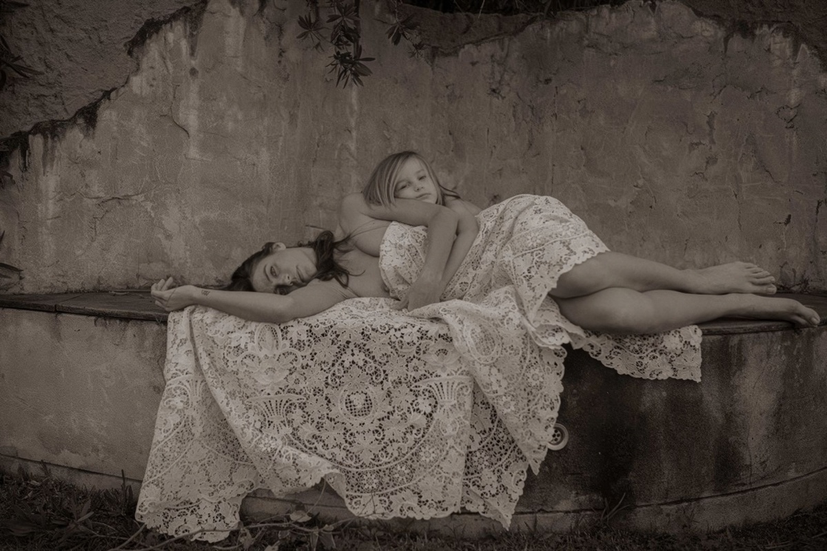 (На фотографии: женщина и ребенок лежат на каменной скамье, накрытые кружевной тканью. Фон — потрескавшаяся стена, создающая контраст между нежностью и суровостью окружающего мира.)