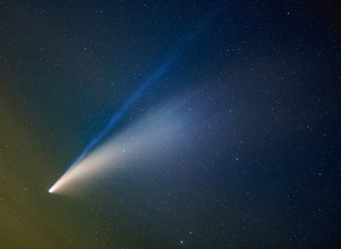 На фото ретроградная комета C/2020 F3, обнаруженная 27 марта 2020 года космическим инфракрасным телескопом NEOWISE. В северном полушарии комету можно было наблюдать невооружённым глазом на протяжении нескольких недель (июль 2020 года). Первый хвост (голубоватый) светится за счёт флуоресценции молекул С2 и CN, а второй хвост состоит из пыли. Наличие двух хвостов получается из-за солнечного ветра. Первый хвост направлен точно от Солнца. А на пыль солнечный свет не может так воздействовать и сильно отклонять её от движения по орбите кометы. Следующий раз комета вернётся к Солнцу через 6800 лет.