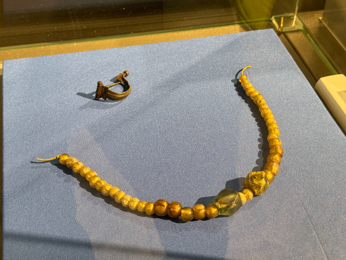 Бусины золотостеклянные, вторая половина I – первая половина II вв н.э., стекло, фольга, формовка. Низка из 41 бусины. Большинство бусин – боченковидные. 27 из них имеют диаметр около 0,9 см. Две крупные бусины – 14-гранные светло-зеленого цвета и желтого стекла.