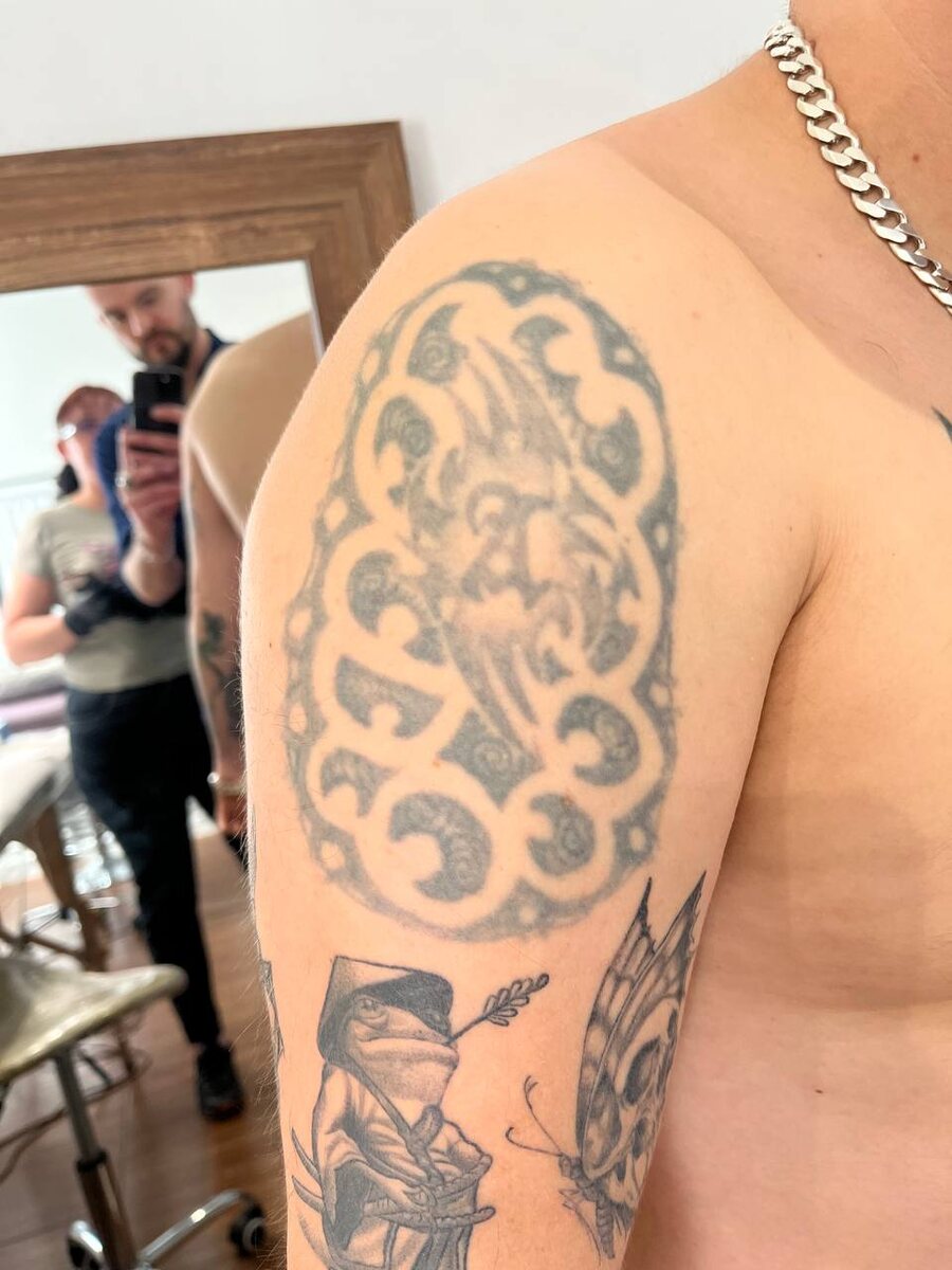 
Первая фотография показывает исходное тату на плече. Оно выполнено в стиле старой школы с орнаментальным узором и изображением, которое со временем сильно поблекло.